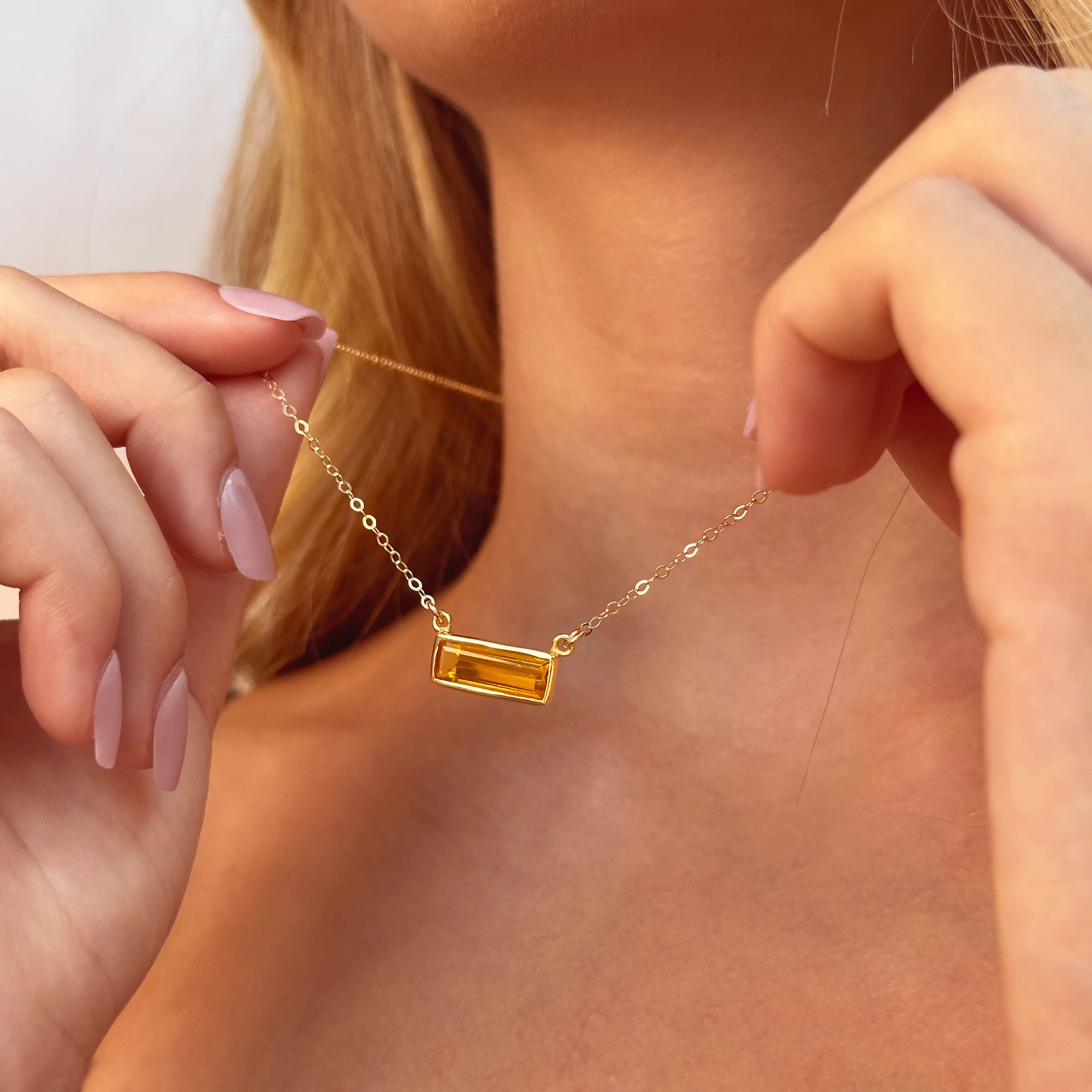 citrine baguette necklace shown on model holding the delicate gold filled chain and vertical bar pendant, sparkling yellow citrine gemstone catching sunlight, personalized engraving available on the bar pendant, dainty November birthstone necklace ideal for women who love minimalist jewelry, elegant layering piece and thoughtful personalized gift for her birthday, graduation or holiday season presented in premium packaging