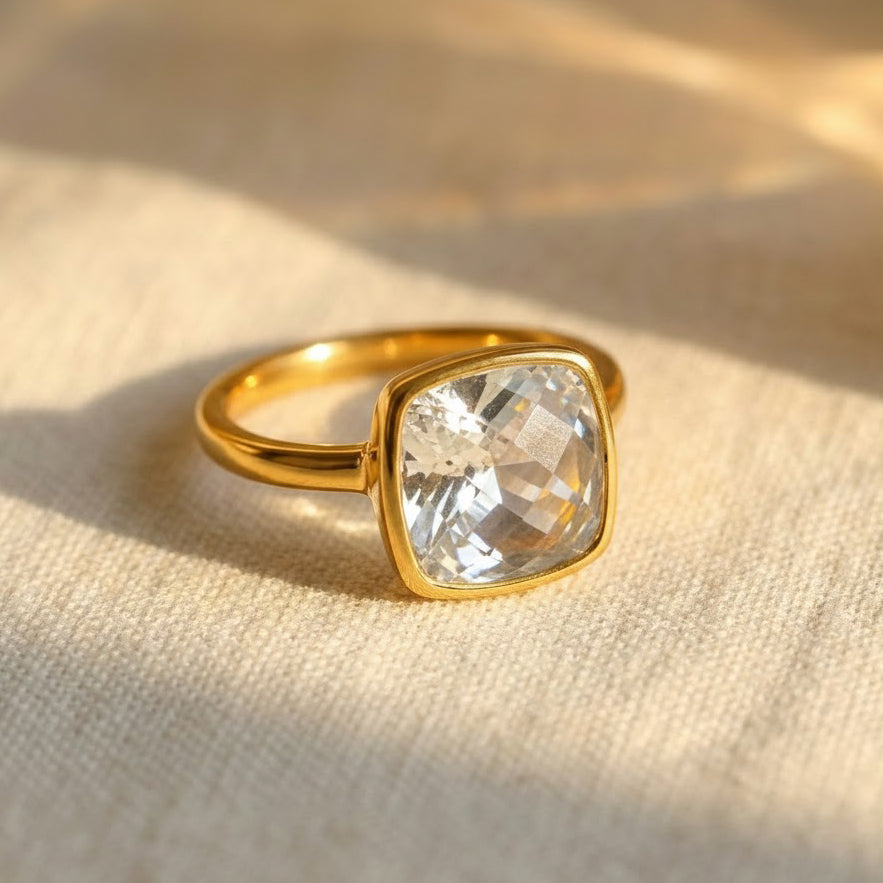 Cushion cut clear quartz ring in polished yellow gold bezel setting resting on soft neutral linen fabric, featuring a 9mm square transparent gemstone with diamond-like brilliance, April birthstone ring for women, minimalist modern fine jewelry, gold bezel set quartz ring, elegant stacking gemstone ring, handcrafted artisan birthstone jewelry, square clear gemstone ring gift for her