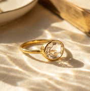 Close-up of oval clear quartz ring in gold on warm neutral fabric, faceted crystal statement ring