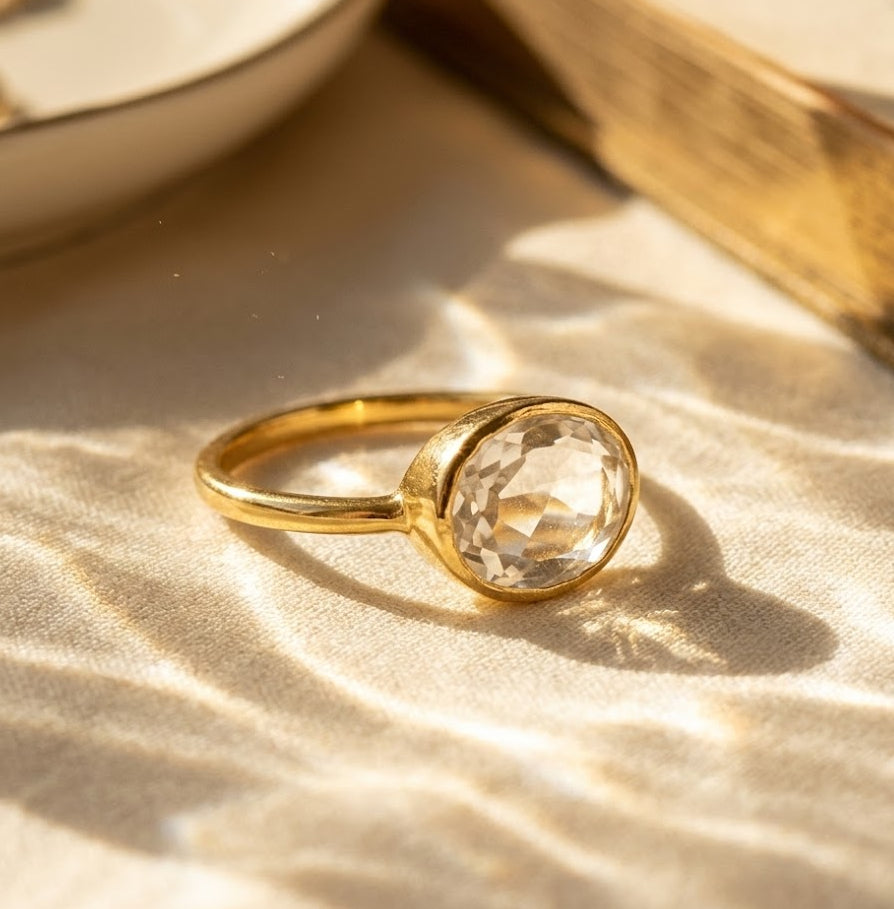 Close-up of oval clear quartz ring in gold on warm neutral fabric, faceted crystal statement ring