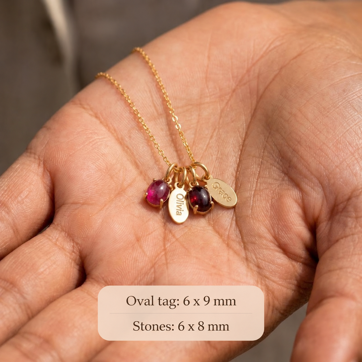 Hand holding personalized oval birthstone and engraved name charms showing petite gemstone and tag sizes.
