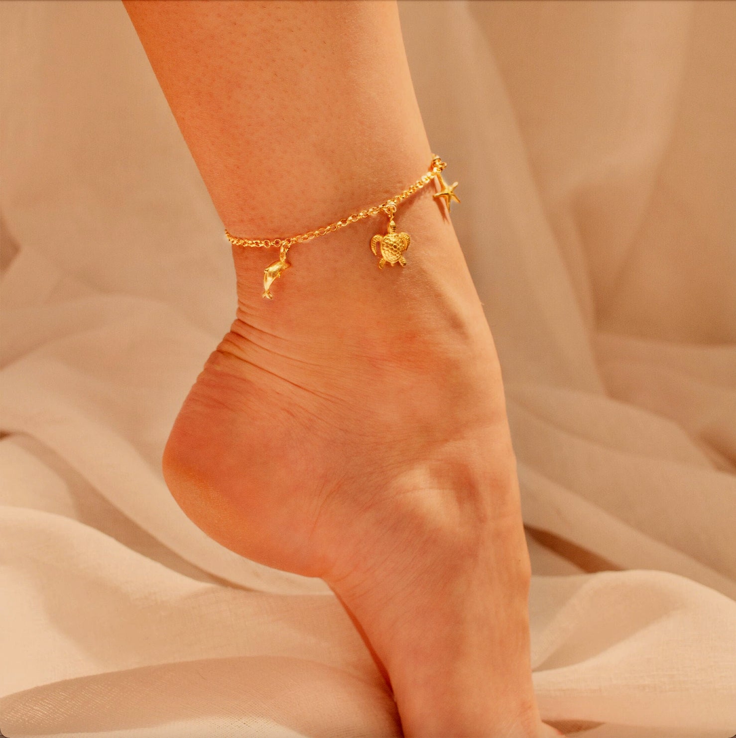 Personalized gold charm anklet worn on ankle against soft neutral fabric, dainty gold filled anklet with symbolic charms, handmade adjustable ankle bracelet for women, minimalist charm jewelry designed for everyday wear or summer styling, lightweight anklet with delicate chain and meaningful charms, custom jewelry gift perfect for birthdays, vacations, or special occasions
