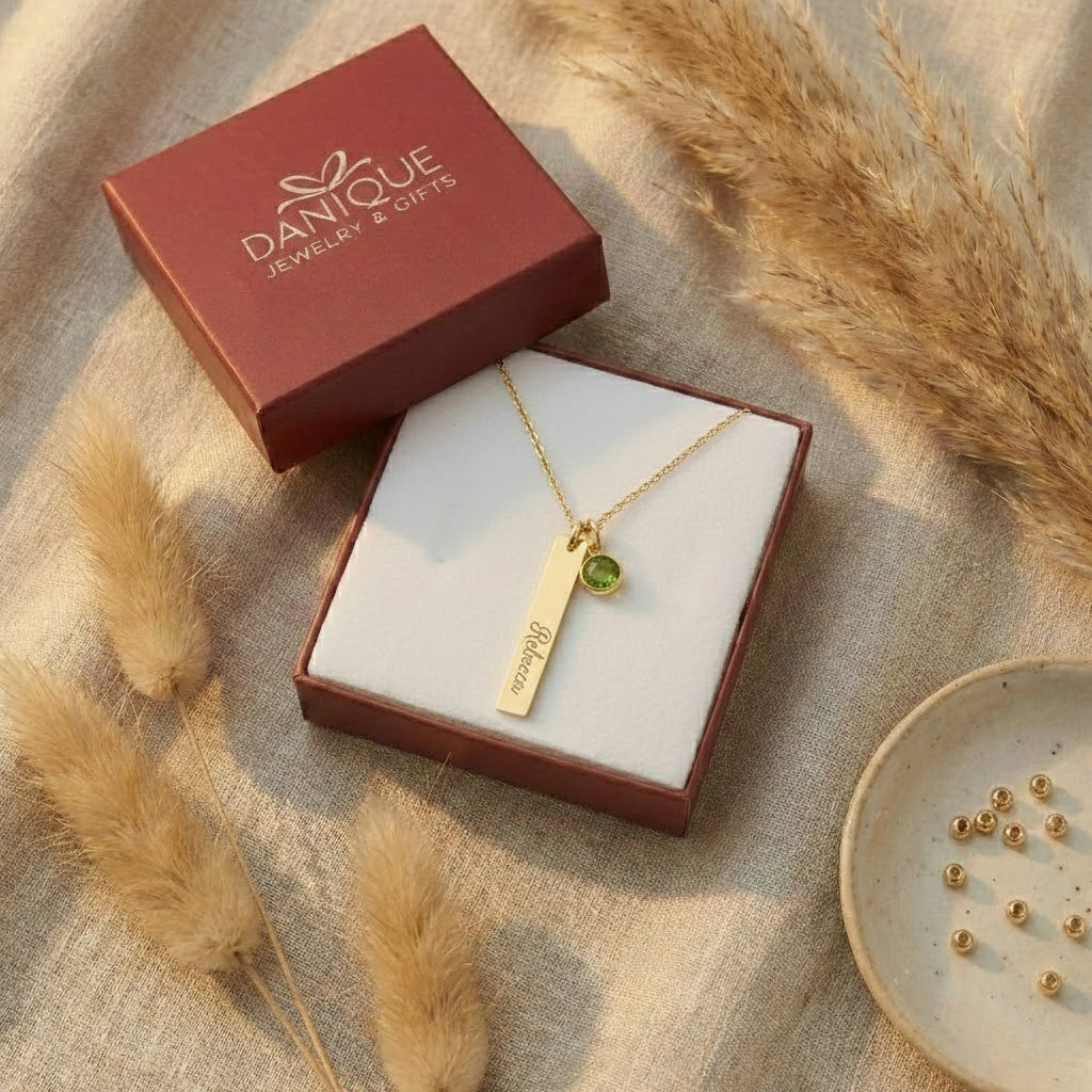 Personalized gold bar and green birthstone necklace presented inside a burgundy Danique Jewelry gift box.