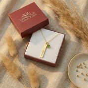 Personalized gold bar and green birthstone necklace presented inside a burgundy Danique Jewelry gift box.