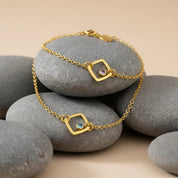 Dainty custom birthstone bracelet with minimalist open square charms resting on smooth gray river stones, each geometric gold link set with a tiny sparkling gemstone that can be personalized with your choice of birthstone. Delicate adjustable chain bracelet in gold, perfect for everyday wear, stacking with other bangles, or gifting to mom, daughter, sister or best friend for birthdays, Mother’s Day, Christmas or anniversaries.