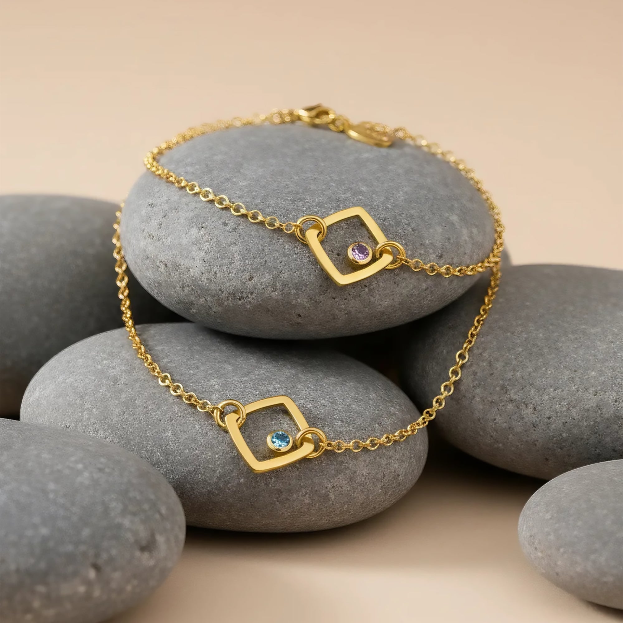 Dainty custom birthstone bracelet with minimalist open square charms resting on smooth gray river stones, each geometric gold link set with a tiny sparkling gemstone that can be personalized with your choice of birthstone. Delicate adjustable chain bracelet in gold, perfect for everyday wear, stacking with other bangles, or gifting to mom, daughter, sister or best friend for birthdays, Mother’s Day, Christmas or anniversaries.