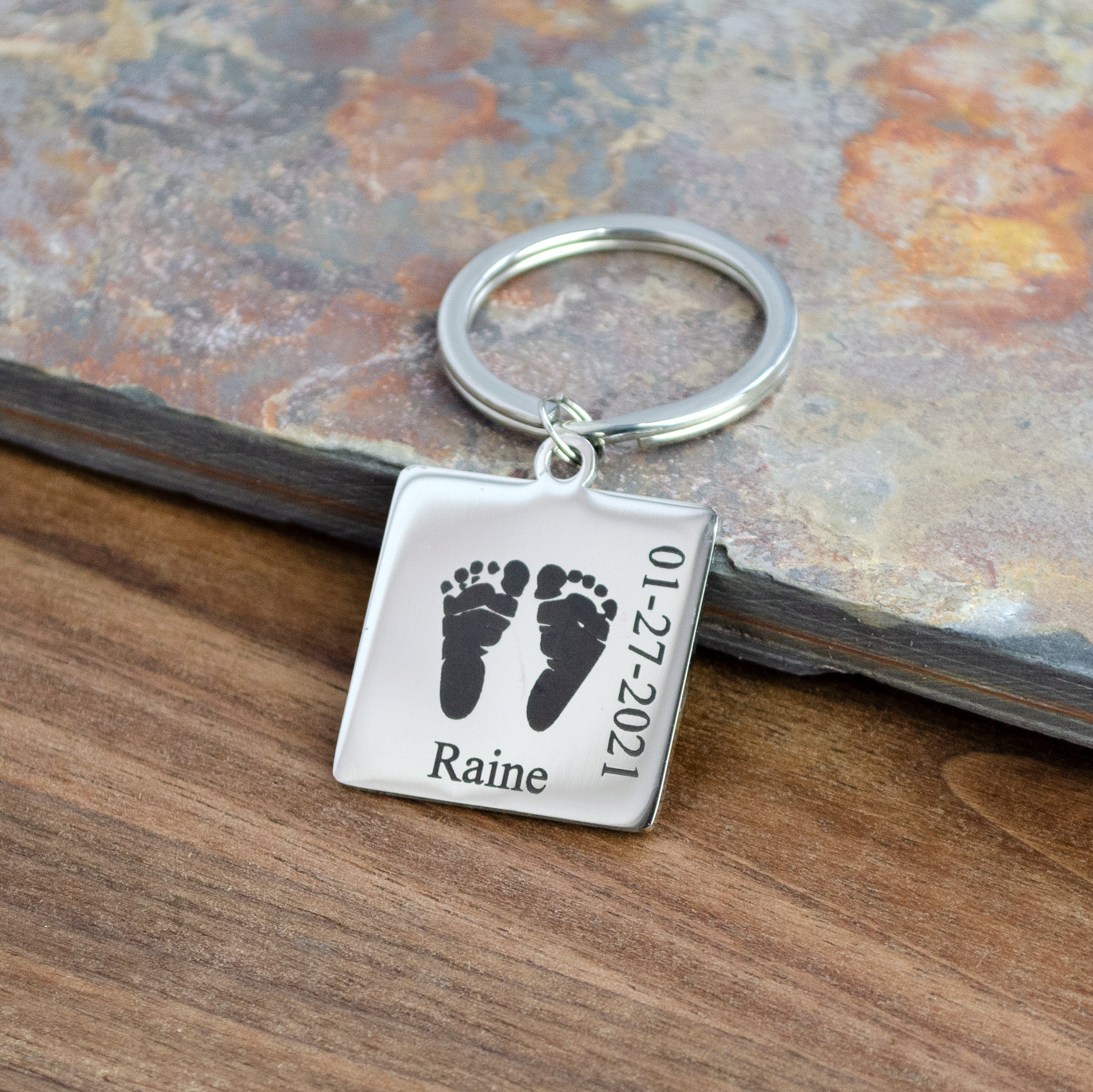 Square Stainless Steel Keychain Custom Engraved with Baby Handprint, Name, and Birth Date Resting on a gray stone tile and wood. 