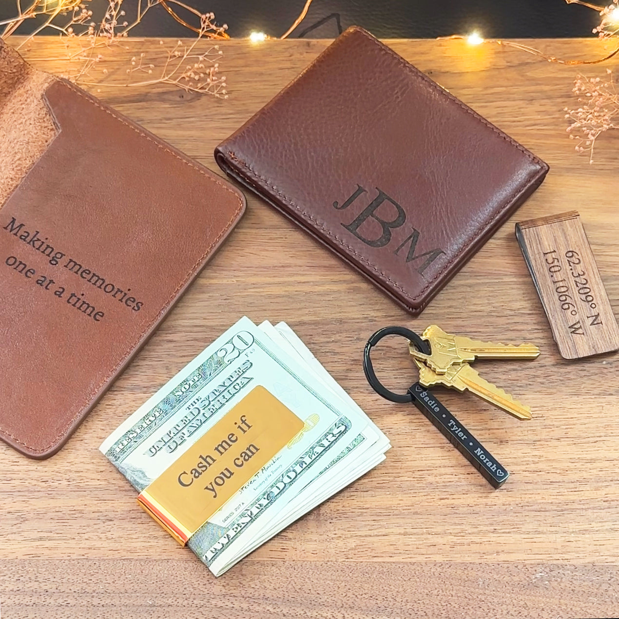 A photo of an open espresso passport holder with inside engraving, a dark brown leather wallet, an engraved coordinates money clip, a gold engraved money clip with cash, and a 3d bar keychain with keys attached and names engraved all on a wood background. 