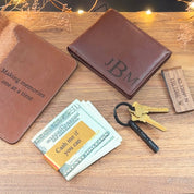 A photo of an open espresso passport holder with inside engraving, a dark brown leather wallet, an engraved coordinates money clip, a gold engraved money clip with cash, and a 3d bar keychain with keys attached and names engraved all on a wood background. 