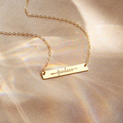 Dainty gold bar necklace engraved with the word “fearless” and a small arrow, laying on a soft champagne satin background with subtle rainbow reflections. Minimalist inspirational word necklace in gold filled finish, perfect motivational gift for friend, daughter or graduate and everyday layering jewelry.