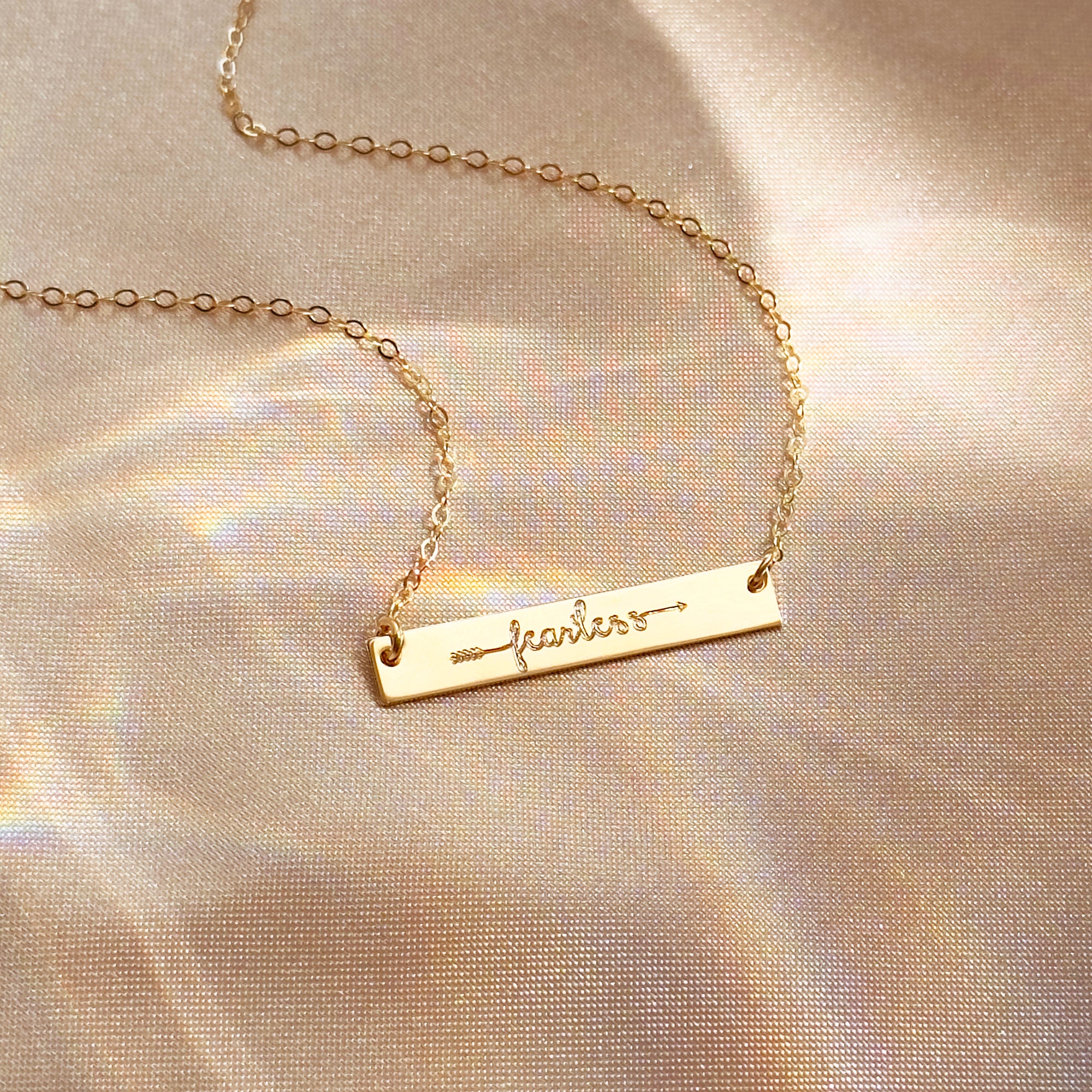 Dainty gold bar necklace engraved with the word “fearless” and a small arrow, laying on a soft champagne satin background with subtle rainbow reflections. Minimalist inspirational word necklace in gold filled finish, perfect motivational gift for friend, daughter or graduate and everyday layering jewelry.