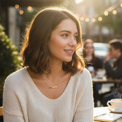Young woman sitting at an outdoor café wearing a dainty gold “fearless” bar necklace, styled with a cozy cream sweater in warm sunset light. Minimalist inspirational word necklace, perfect everyday jewelry gift for friend, daughter, graduate or self-love reminder.