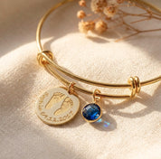 Close-up of custom baby footprint bangle with engraved name, date, and blue birthstone charm on gold adjustable bracelet.