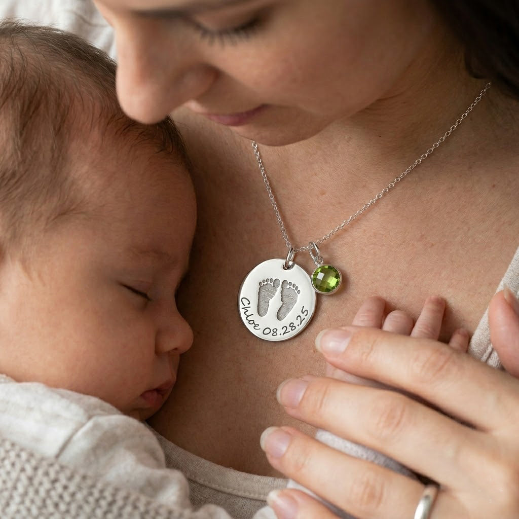 Mother wearing personalized baby footprint necklace with birthstone charm while holding a sleeping newborn.