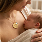 Mother holding newborn while wearing personalized baby footprint necklace with engraved name, date, and birthstone charm.
