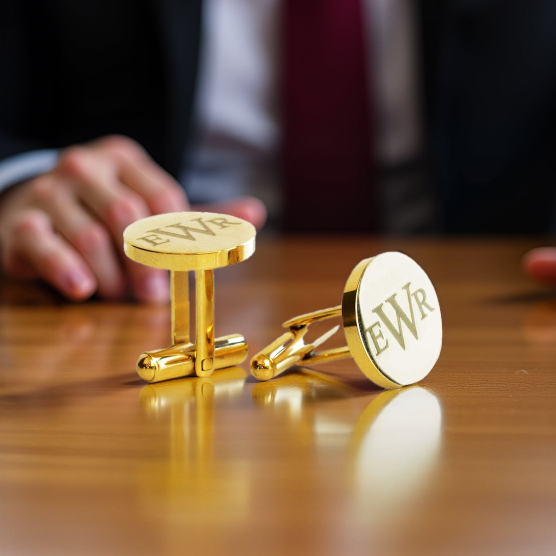 Close-up of personalized gold metal cufflinks engraved with monogram initials "EWR," resting on a polished wooden surface with a blurred suited figure in the background. These custom round cuff links are ideal gifts for weddings, groomsmen, father of the groom, father of the bride, best man, or anniversary. Made from high-shine gold-plated metal, they offer a timeless, elegant touch for formalwear. Perfect men’s accessories for special occasions, personalized gift for him, or custom engraved jewelry.