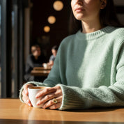 Woman wearing a small oval green amethyst ring while holding a coffee cup at a café, featuring a faceted green amethyst gemstone set in a smooth bezel on a delicate band, styled for everyday wear. Minimalist February birthstone ring shown on hand in natural light, highlighting the soft green hue of the amethyst, elegant oval shape, and timeless design. Dainty gemstone ring perfect for stacking, gifting, or daily wear in sterling silver or gold vermeil, photographed in a cozy coffee shop setting for lifestyl