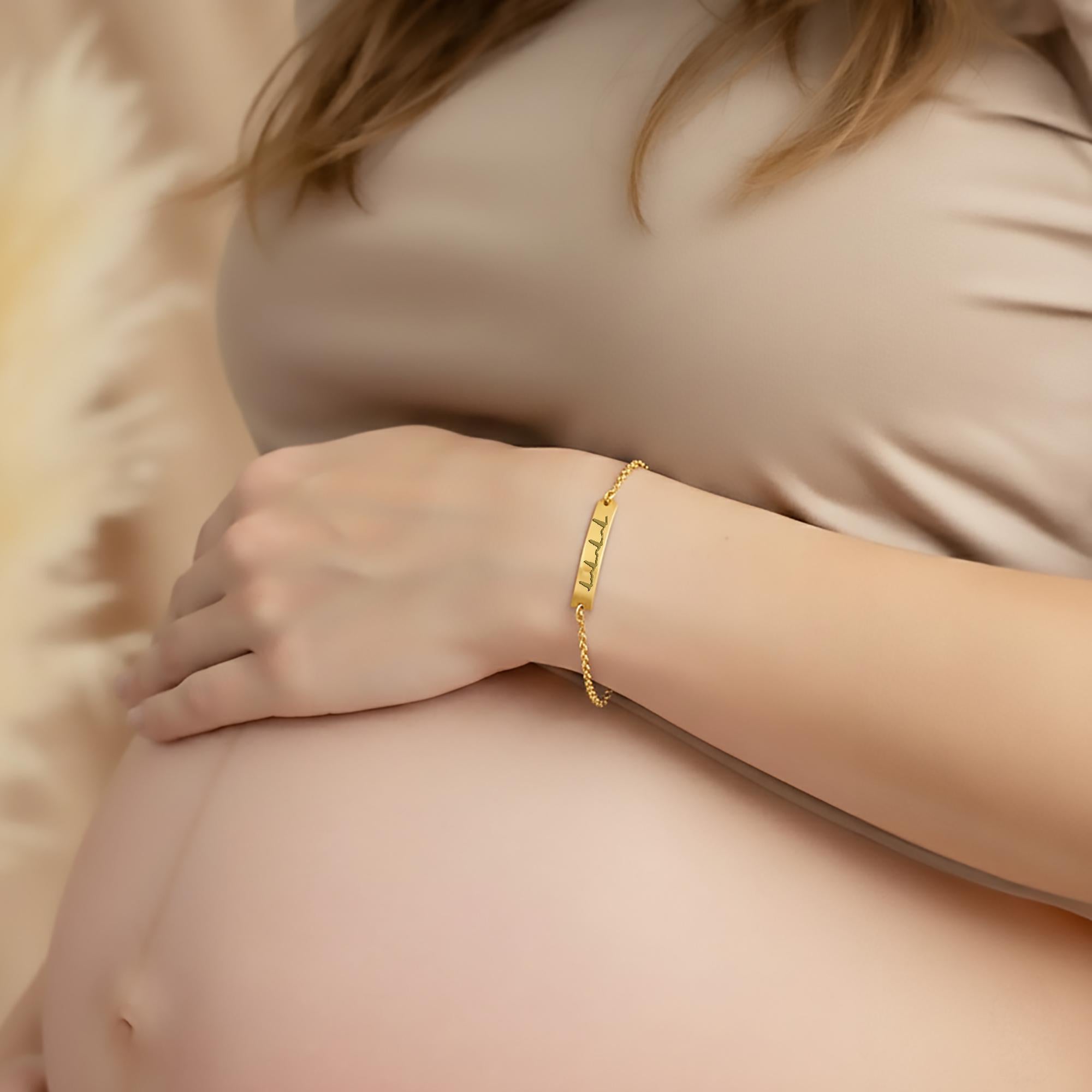 Custom gold bar bracelet with engraved heartbeat design worn by an expecting mother resting her hand on her pregnant belly, meaningful pregnancy jewelry symbolizing baby’s heartbeat; elegant personalized bracelet for maternity photoshoot, baby shower gift, mama-to-be present, sentimental keepsake jewelry for new mom, delicate gold chain accessory with minimalist engraved medical heartbeat motif.