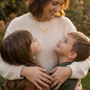 Mother hugging her two children while wearing a gold mama bird necklace with baby bird charms. Meaningful family jewelry symbolizing motherhood, love, and children. Personalized necklace gift for mom.