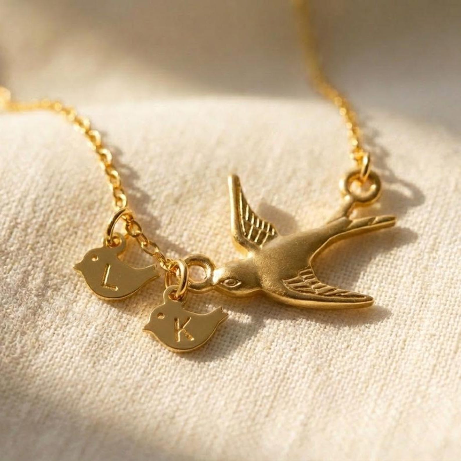 Gold mama bird necklace with two tiny baby bird charms engraved with initials L and K, resting on soft ivory fabric in warm sunlight. Personalized family necklace symbolizing motherhood, children, and love.