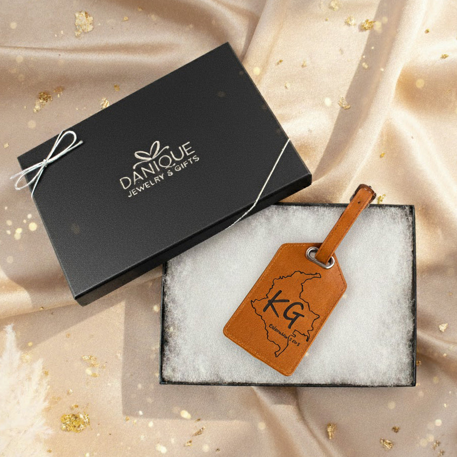 Custom engraved leather luggage tag displayed in a gift box with Danique Jewelry & Gifts packaging, featuring personalized initials “KG” and a custom map outline; warm holiday background highlights this handcrafted travel accessory, perfect for travelers, weddings, and meaningful personalized gifts.