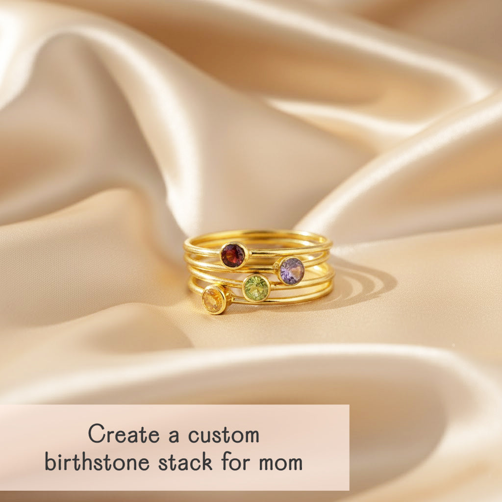stack of dainty gold birthstone rings arranged in a tower on soft champagne satin, each slim band featuring a bezel set gemstone in warm red, lavender, fresh green and golden yellow, casting a gentle shadow in the light, caption reading “create a custom birthstone stack for mom,” showcasing how customers can choose different stones to represent each child, perfect personalized mother’s ring set or meaningful Mother’s Day, birthday or Christmas gift for her