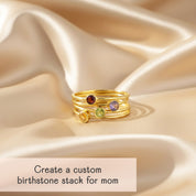 stack of dainty gold birthstone rings arranged in a tower on soft champagne satin, each slim band featuring a bezel set gemstone in warm red, lavender, fresh green and golden yellow, casting a gentle shadow in the light, caption reading “create a custom birthstone stack for mom,” showcasing how customers can choose different stones to represent each child, perfect personalized mother’s ring set or meaningful Mother’s Day, birthday or Christmas gift for her