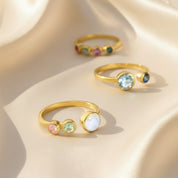 Multi birthstone adjustable rings for mom displayed on soft champagne satin, each handcrafted in warm gold with bezel-set gemstones. Front ring shows the classic mom of two design with one large glowing moonstone 5mm center stone and two smaller pastel child birthstones, while the others feature different family stone combinations. These personalized family rings make meaningful Mother’s Day, birthday or Christmas gifts, celebrating children with colorful, dainty everyday jewelry.