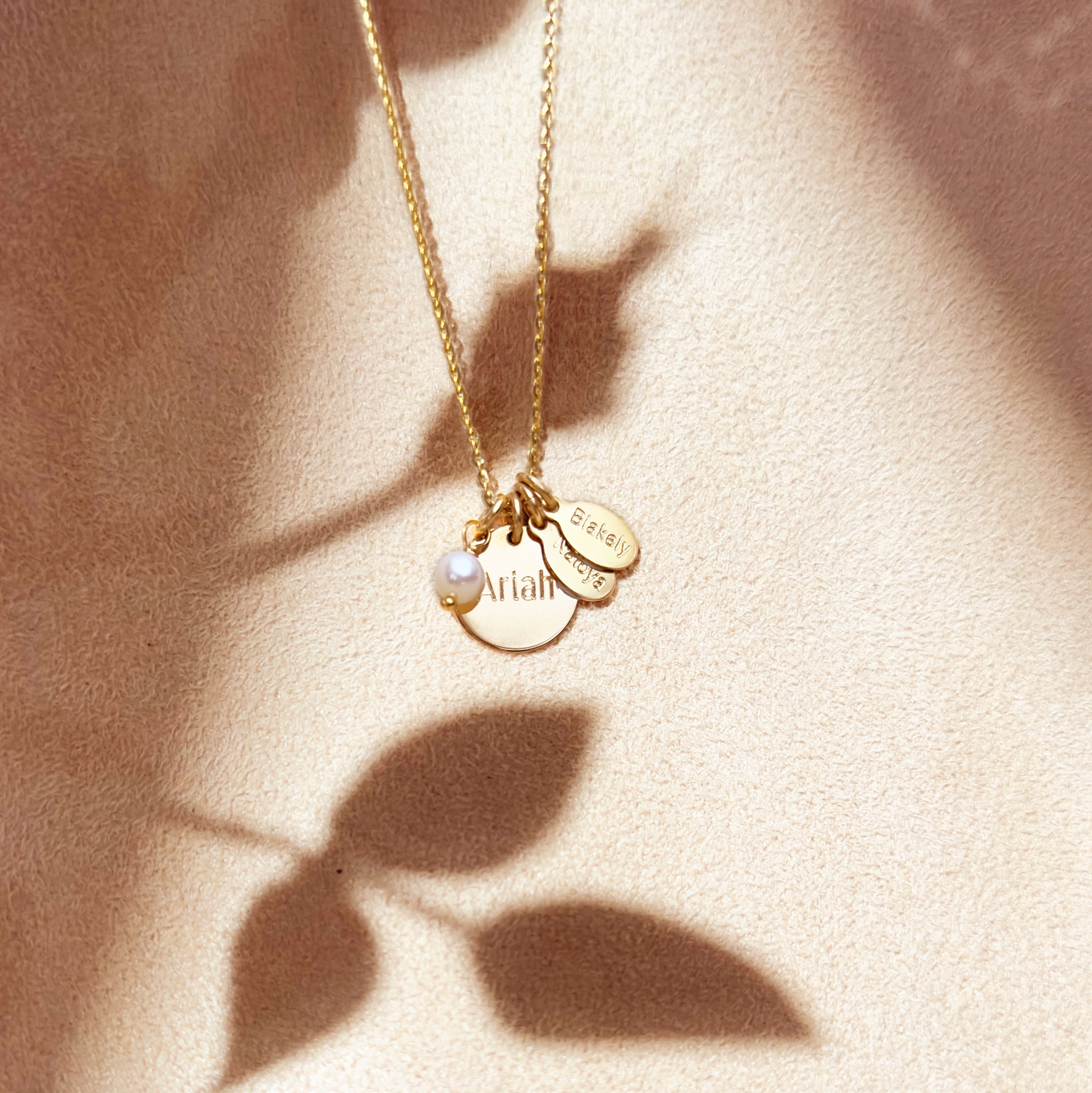 Gold disc and custom name tag necklace on beige background. Engraved with Ariah on circle disc. Elegant and timeless, personalized Mother's necklace features a round disc with your name, oval tags for your kids and a freshwater pearl charm. Mom necklace, kids names, pearl charm.