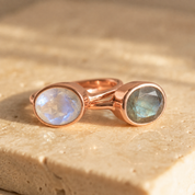 Rose-gold-vermeil moonstone and labradorite oval rings displayed together on a beige stone surface.