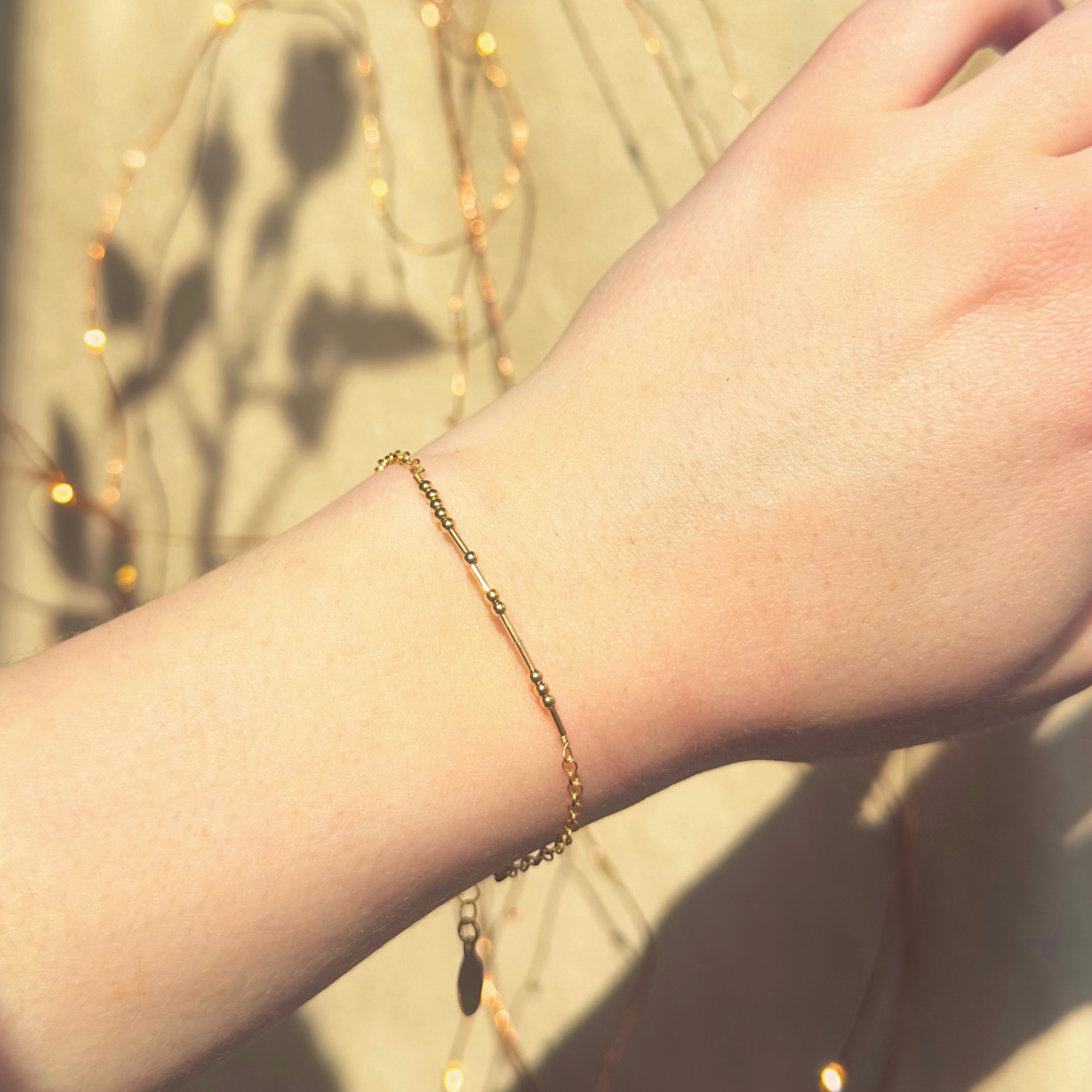 A light-skinned person wears a gold beaded morse code bracelet. The shadow of the person's hand in the bottom right. The shadow of a rose is in the upper left. Gold bracelet, beaded bracelet, personalized bracelet, secret code bracelet, personalized gold jewelry for girls, trendy gold bracelets for women, jewelry trends for teenage girls and young women