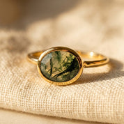 Oval moss agate ring in gold vermeil displayed on soft beige fabric, showing the faceted green gemstone and delicate stacking band.