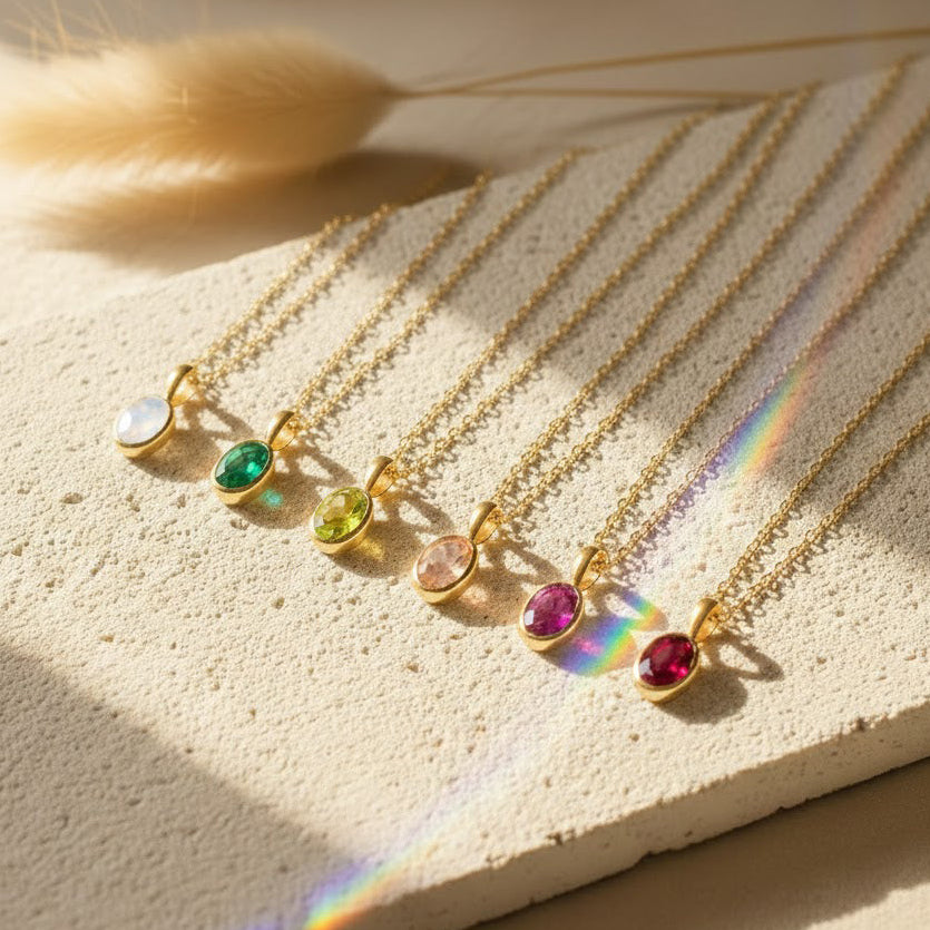 Oval cut birthstone bezel pendant necklaces displayed on textured neutral stone, featuring multiple gemstone colors representing different birth months. Dainty gold birthstone necklaces arranged in a row, highlighting minimalist bezel set design and delicate chain. Personalized birthstone jewelry perfect as a custom gift for her, Valentine’s Day gift, mom necklace, or meaningful everyday gemstone pendant.