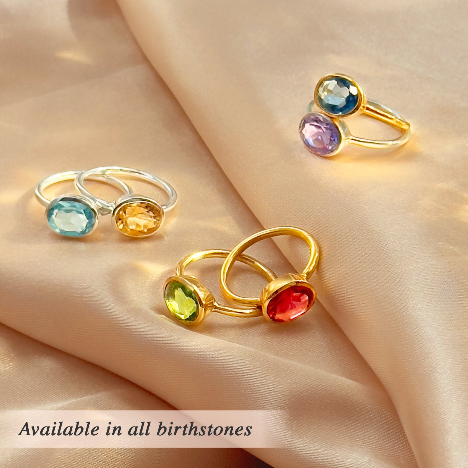 Assorted oval birthstone rings arranged on blush satin—bezel-set ovals on yellow-gold tone and polished sterling-silver bands. Build a colorful ring stack or bold statement ring look with gemstones such as aquamarine (March), citrine (November), peridot (August), ruby red (July), and amethyst (February). A modern collection of cocktail rings for everyday style or anniversary jewelry for her.