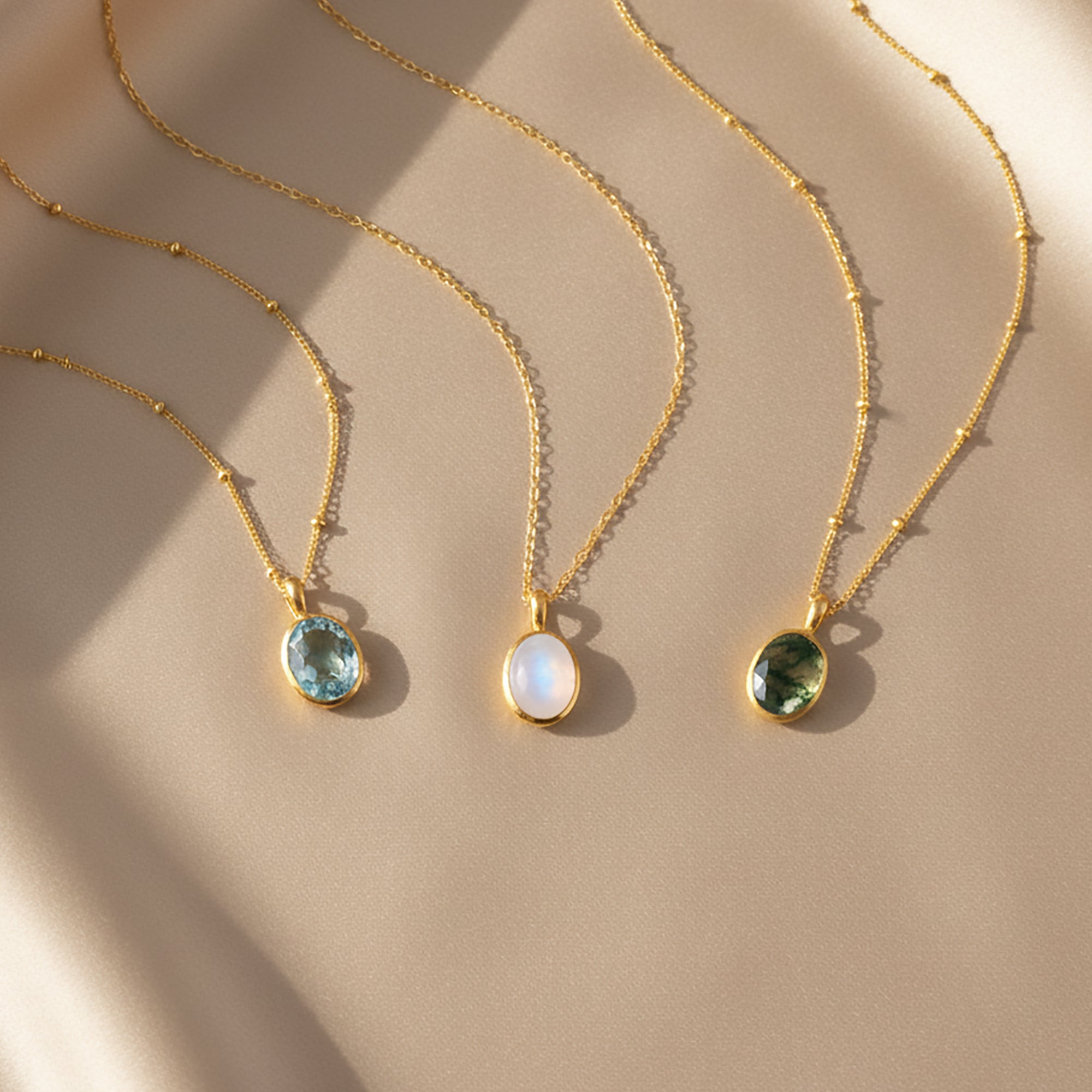 Dainty oval gemstone necklaces displayed on a soft beige background in warm sunlight, featuring three bezel-set stones in blue, iridescent moonstone white and deep green on delicate gold satellite chains. Minimalist birthstone or gemstone pendant necklaces perfect for everyday wear or layering, a meaningful gift for her, mom, sister or best friend for birthdays, Mother’s Day, Christmas, anniversaries or bridesmaid jewelry.