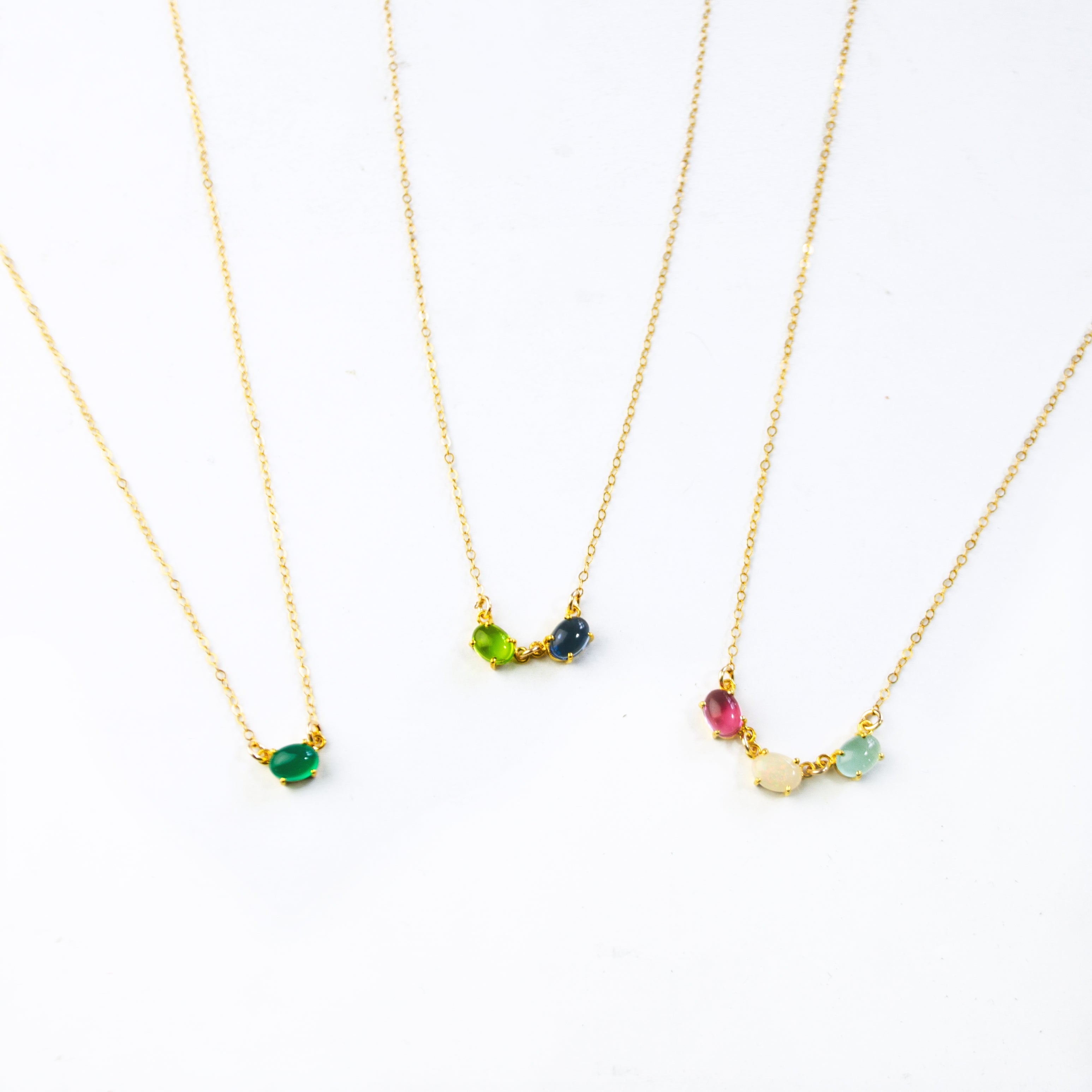 Three gold filled oval prong set cabochon stone necklaces. First necklace features  single green onyx stone. The middle necklace has peridot and kyanite birthstones. Final necklace features ruby, moonstone and aqua chalcedony birthstones. 