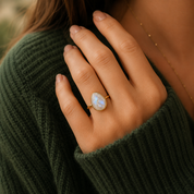 A gold ring with a teardrop-shaped iridescent white stone, worn on a person's ring finger. The person is wearing a dark green ribbed knit sweater, and a delicate gold chain necklace is visible against a warm, softly blurred background.