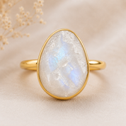 A gold ring featuring a large, teardrop-shaped white moonstone gemstone with a soft blue iridescent sheen, set against a soft beige background with delicate dried flowers in the upper left corner.