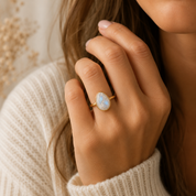 A close-up lifestyle shot of a woman wearing a gold ring with a teardrop-shaped light blue opal gemstone on her finger, set against a soft cream knit sweater and a warm, blurred background.