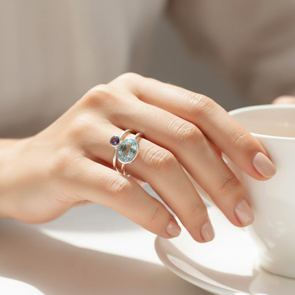 Close-up hand wearing a personalized birthstone stacking ring set, dainty bezel gemstone rings with a larger oval stone for mom and tiny round stones for children, minimalist mothers ring set in gold vermeil or sterling silver, custom family jewelry gift for mom of two, mom of three, or grandma, choose January garnet and March aquamarine combinations by birth month, delicate stacking rings for everyday wear