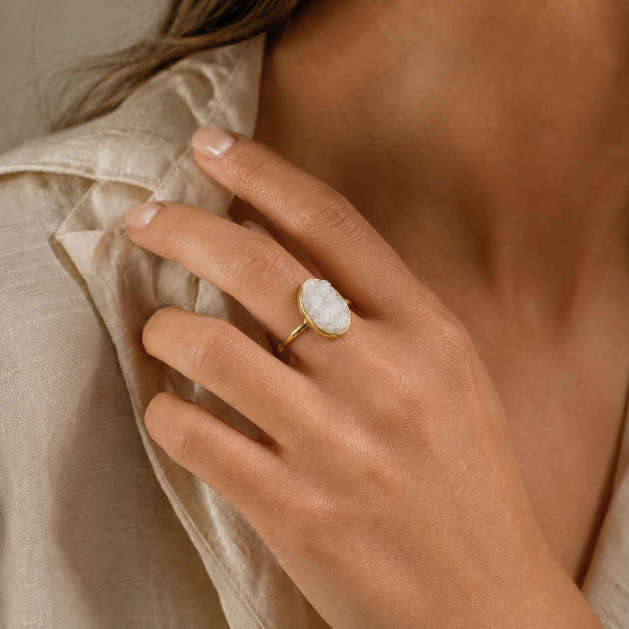 A close-up lifestyle shot of a woman's hand wearing a gold ring with a textured white stone, set against a soft beige linen shirt.