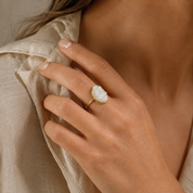 A close-up lifestyle shot of a woman's hand wearing a gold ring with a textured white stone, set against a soft beige linen shirt.