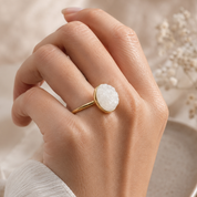 A gold ring with a textured, oval-shaped white stone is shown on a person's hand, set against a soft beige fabric background with dried flowers.