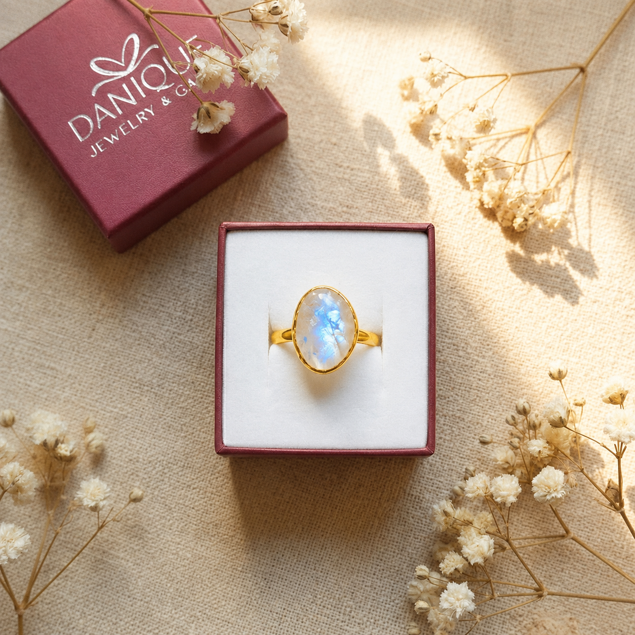 A gold ring with an oval iridescent blue-white stone sits in an open white jewelry box, with a dark red Danique Jewelry & Gifts box and dried white flowers arranged around it on a beige fabric background.