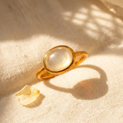 Oval gemstone ring shown on beige background with mother of pearl cabochon center stone and simple minimalist band.