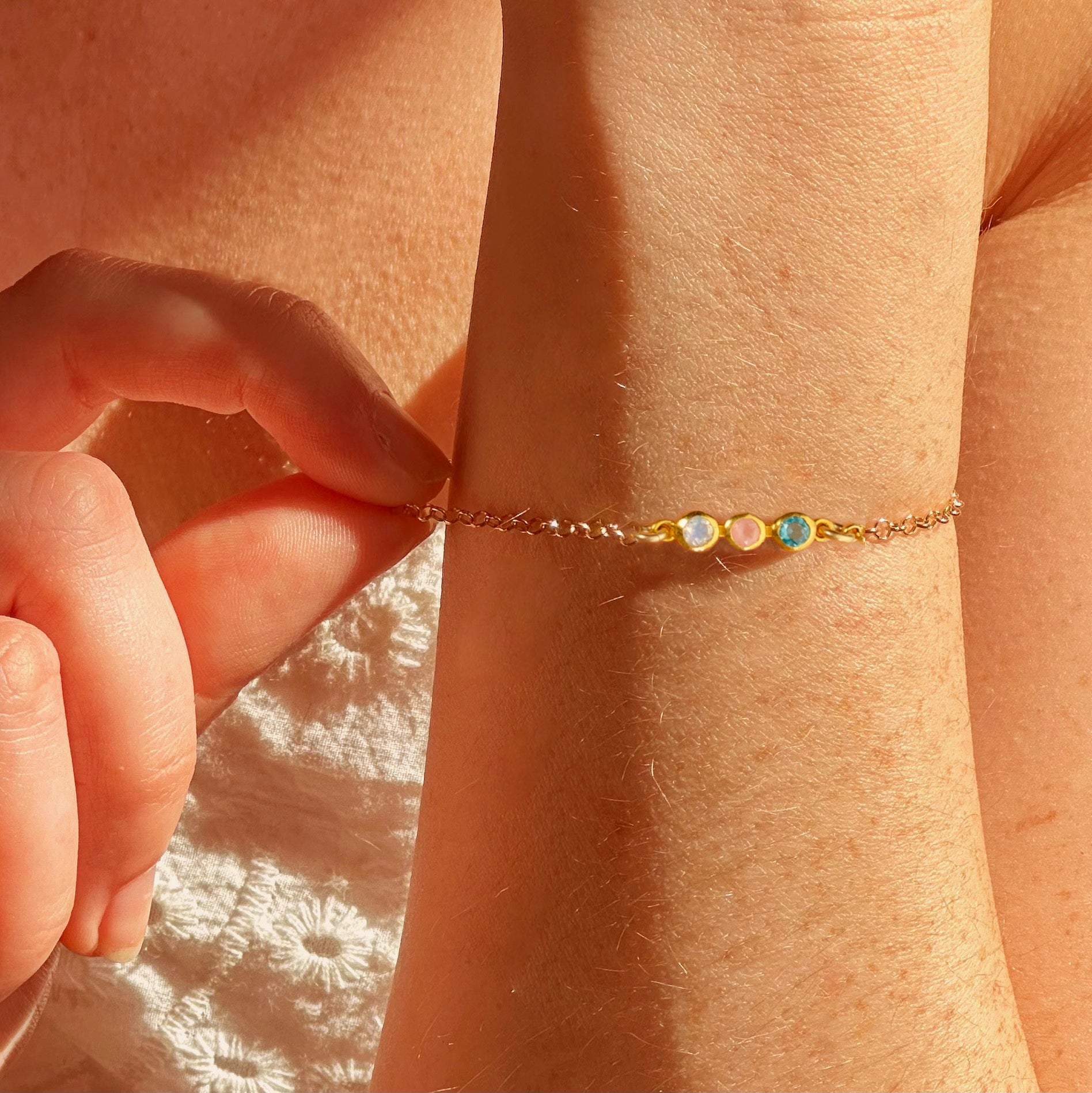 Close-up of tiny gemstone bar bracelet on wrist, crafted in gold filled chain with bezel-set gemstones: june moonstone, december blue topaz, october fire opal This dainty personalized bracelet makes a meaningful jewelry gift for mom, daughter, grandmother, or loved one. Perfect for birthdays, Mother’s Day, anniversaries, or Christmas, it comes in sterling silver, yellow or rose gold filled, with customizable length and gemstone selection for a unique keepsake.
