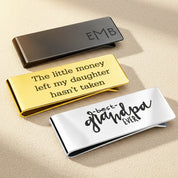 Three polished money clips in gold, black, and silver with custom engravings, displayed on a soft surface with sunlight reflections.