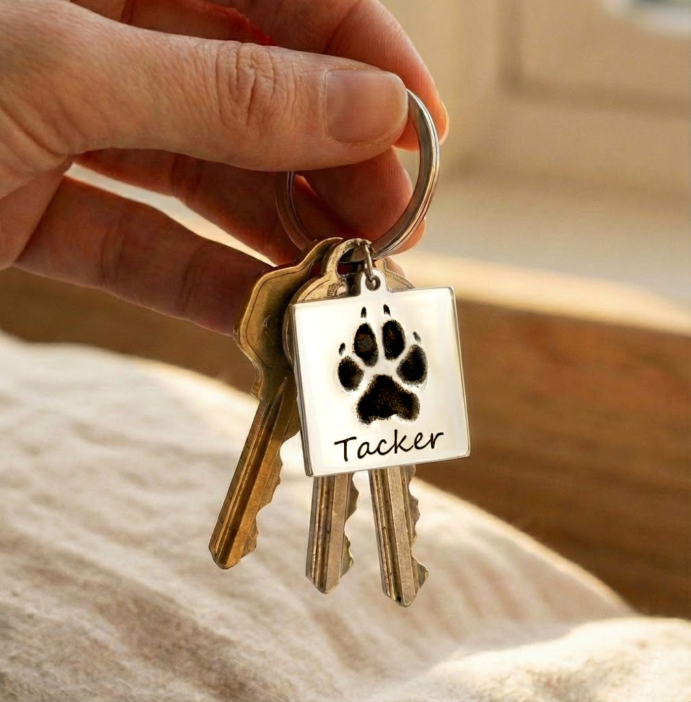 Personalized pet paw print keychain shown attached to a set of house keys, featuring a custom engraved dog paw print and pet name on a square stainless steel charm. This custom pet memorial keychain is handcrafted as a meaningful keepsake for dog and cat owners, pet parents, and animal lovers. Ideal as a sympathy gift, pet loss remembrance, or everyday personalized pet accessory, designed to keep a beloved pet close at all times.