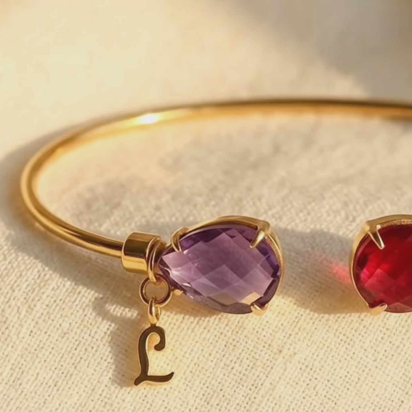 Video showing a personalized birthstone bangle bracelet with two custom faceted gemstones and two initial charms, featuring a purple amethyst and ruby quartz stone in a delicate gold bracelet setting.