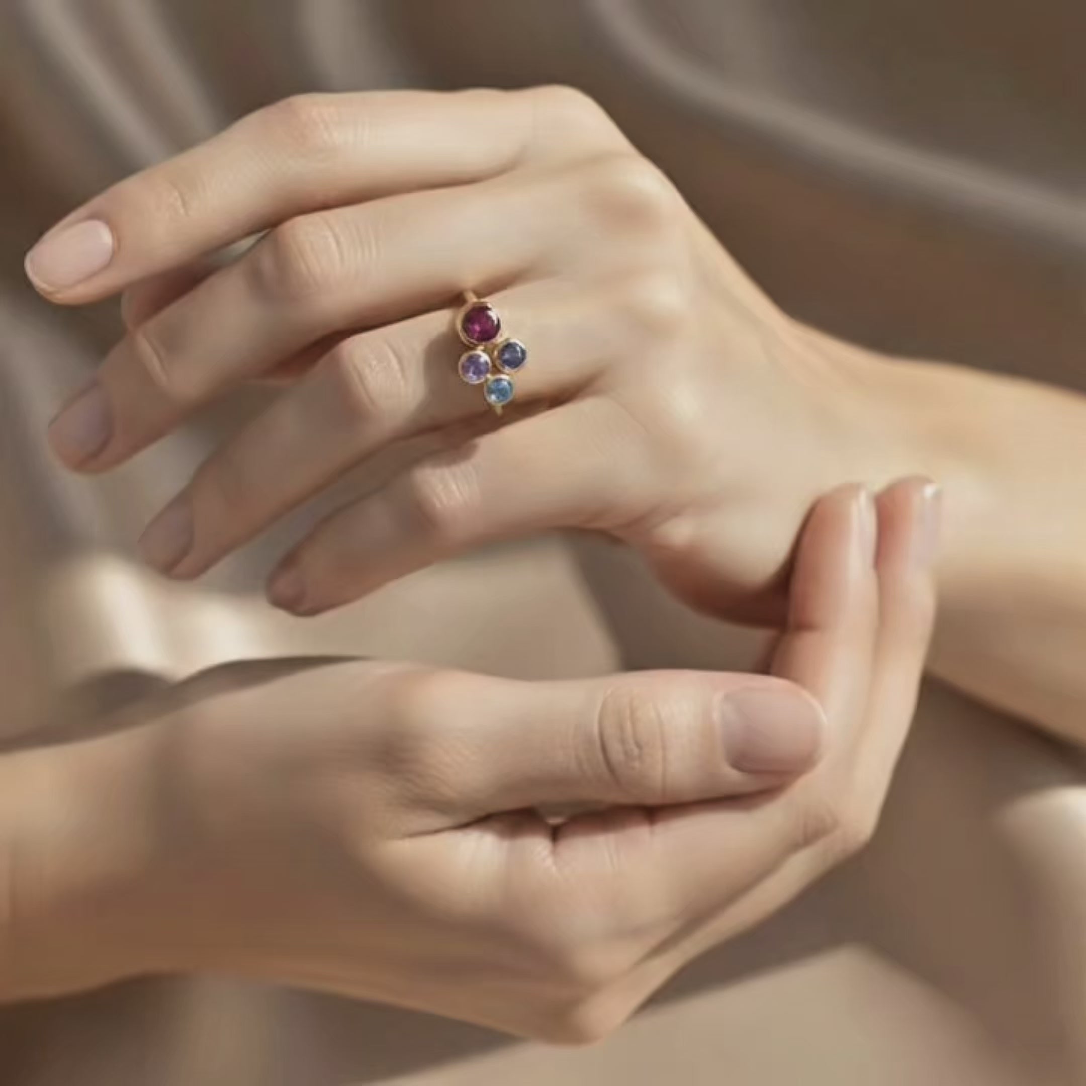 Video showing a close-up of a woman’s hands gently turning a custom birthstone ring for mom, featuring a yellow gold band with four bezel-set multi-colored birthstones symbolizing children or family, sparkling against a soft satin beige background; personalized family ring, dainty stacking style, meaningful Mother’s Day or birthday gift for mom or grandma.